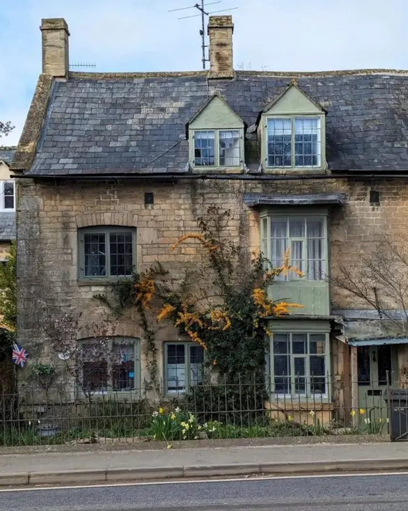 A stone English country house with sage green trim on its dormer and bay windows.