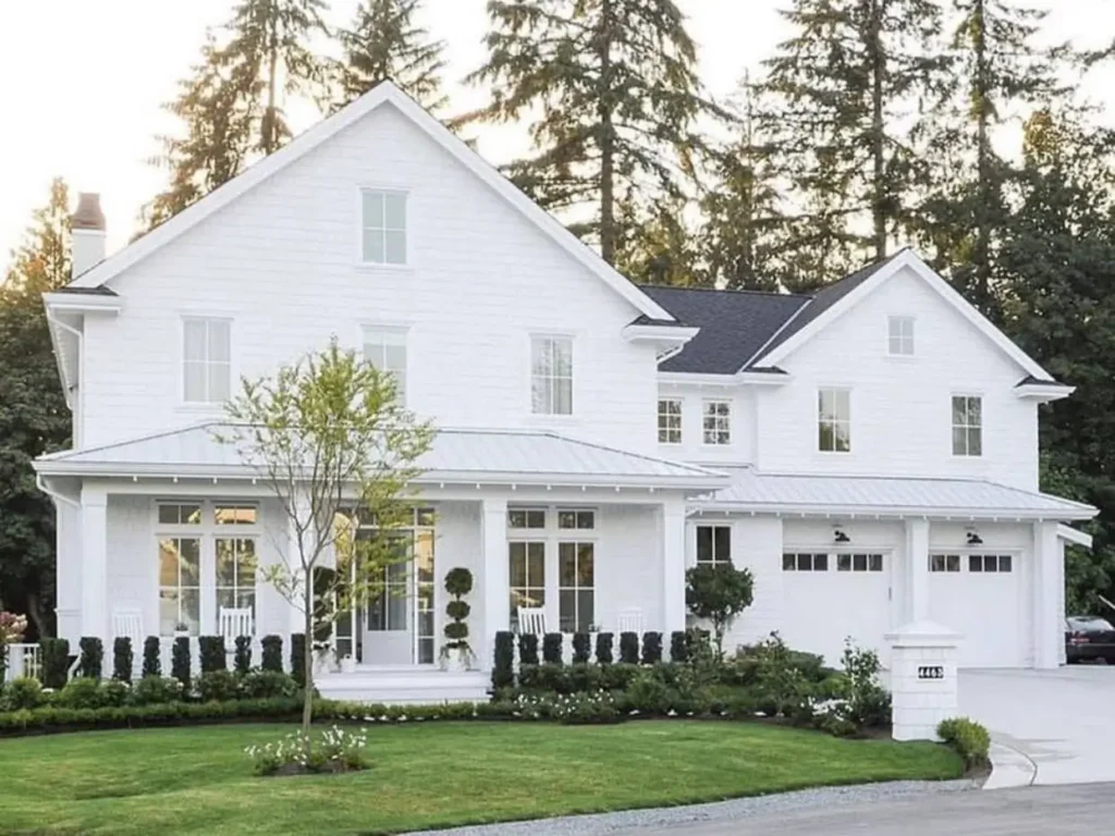 An elegant, large off-white farmhouse with matching white trim and windows, set against a backdrop of tall pine trees.
