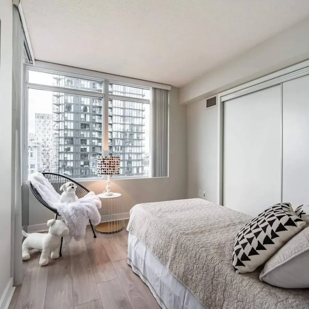 A small, minimalist bedroom with a large window overlooking a city, featuring a modern black accent chair and a bed with a simple quilt.