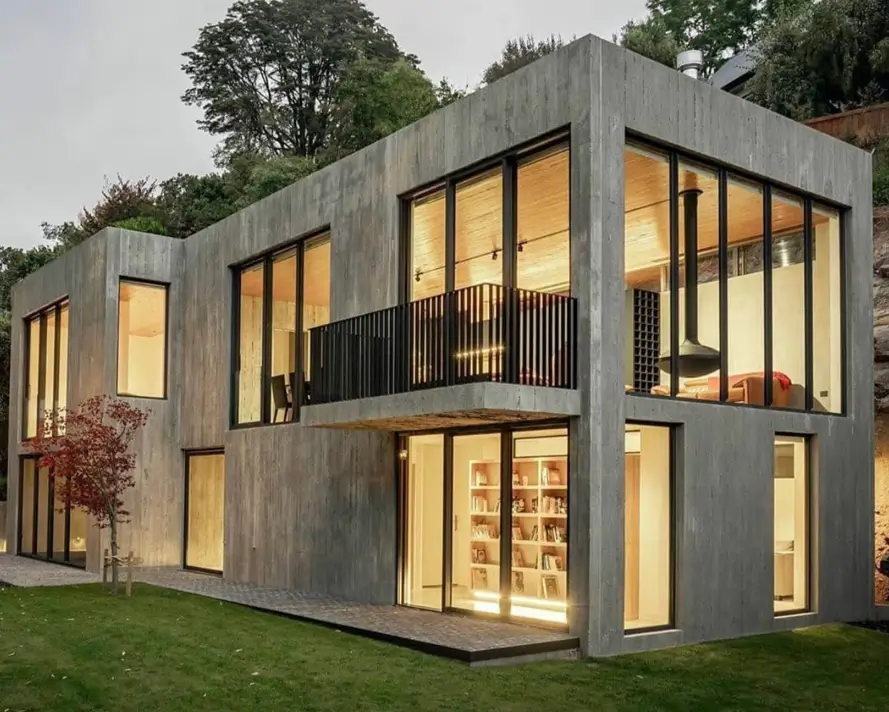 A two-story brutalist house made of board-formed concrete, its minimalist form warmed by large, glowing windows that reveal a cozy interior at dusk.