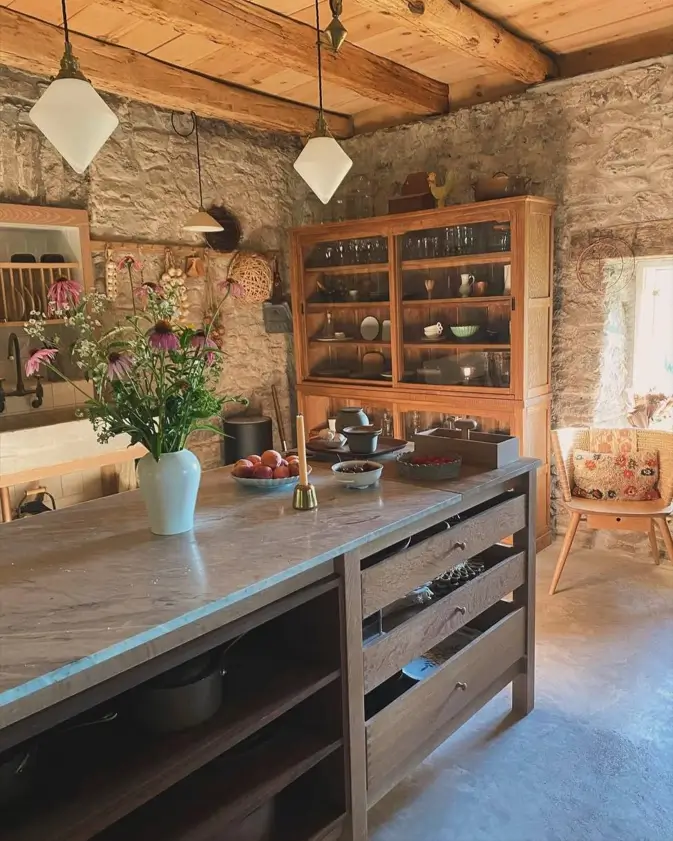 A rustic kitchen featuring exposed stone walls and a large, freestanding wooden hutch displaying glassware.