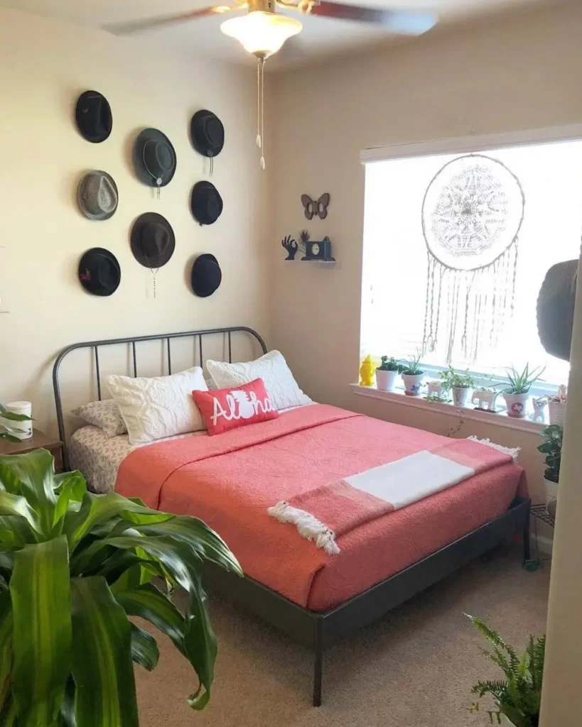 A small cozy bedroom with a collection of hats displayed as art above a metal-frame bed with a coral-colored quilt.