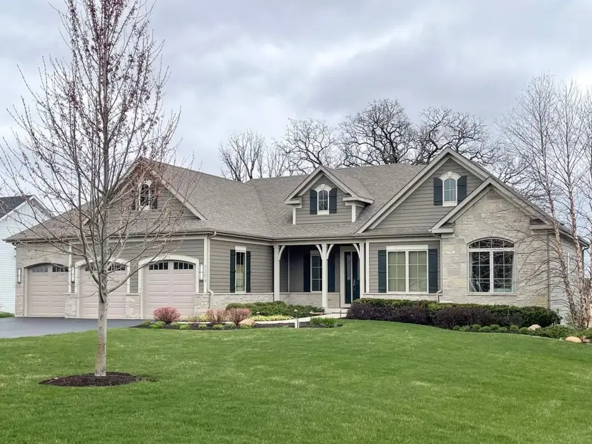 A large traditional ranch house with taupe siding, stone accents, and dark green shutters on an overcast day.