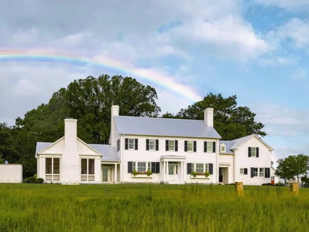 A sprawling historic off-white farmhouse with black shutters and large chimneys, set in a green field with a rainbow overhead.