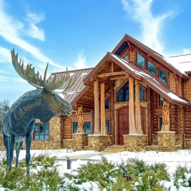 Embracing Grandeur: 25 Inspiring Ideas for Large Log Cabin Homes 25 The grand entrance of a large log cabin with massive log pillars and a bronze moose statue in the snow.