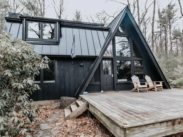 A modern black A-frame cabin with a large natural wood deck extending the living space into the surrounding woods.