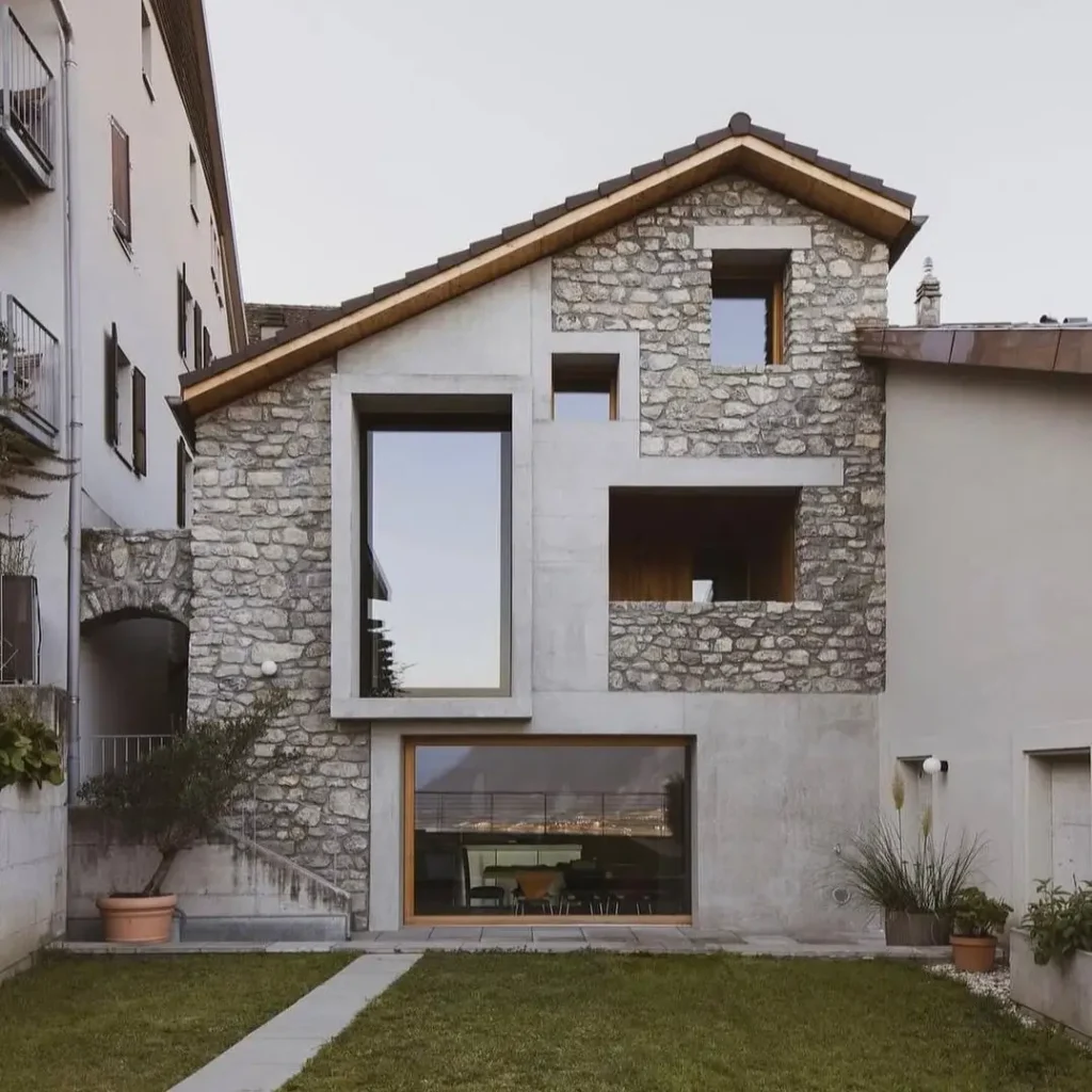 A rustic stone house with modern concrete sections and large geometric windows set into the facade.