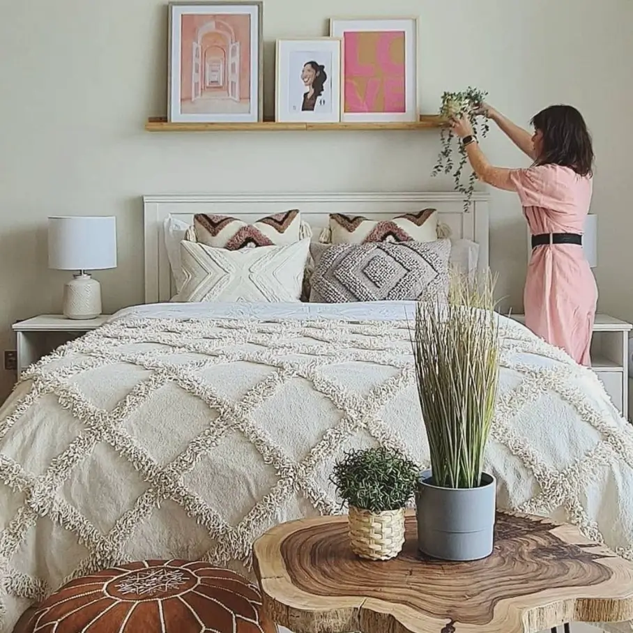 A woman in a pink dress arranges a plant on a wooden art ledge above a bed, which displays a collection of framed art prints.