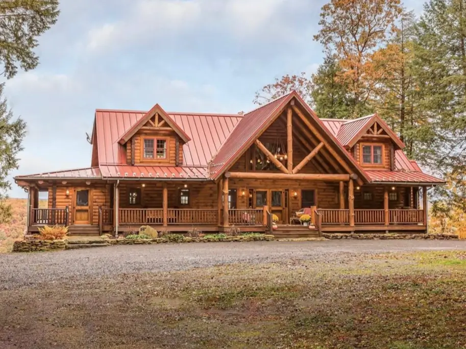 Embracing Grandeur: 25 Inspiring Ideas for Large Log Cabin Homes 26 A classic log cabin with a red metal roof and a full wraparound porch, set against a backdrop of autumn trees.