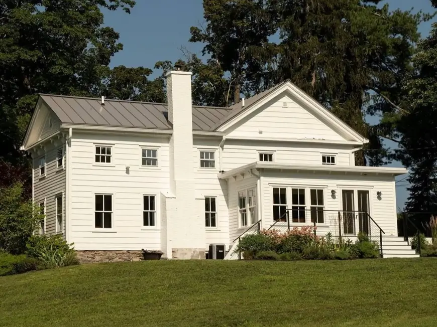 The rear of an off-white farmhouse on a hill, featuring a prominent painted brick chimney and stone foundation.