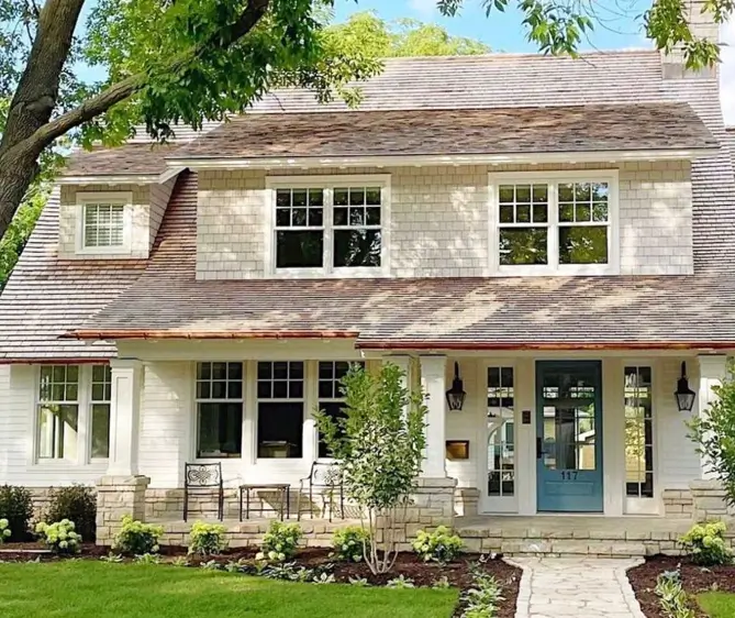 A modern country house design featuring mixed materials like cedar shakes, white siding, and stone pillar bases.