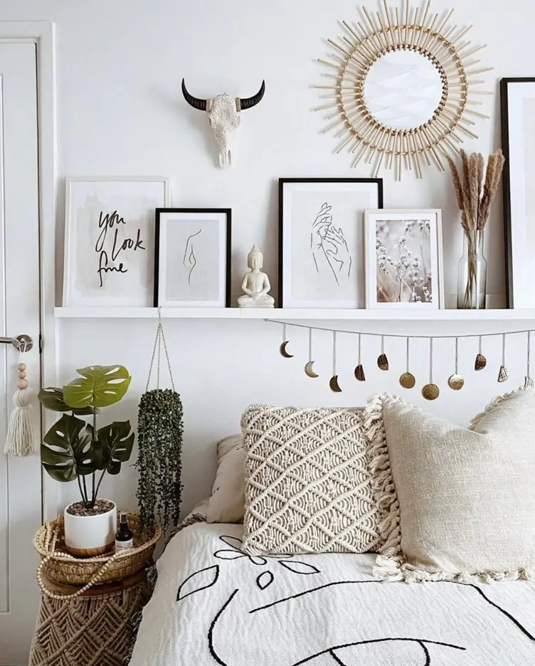 A white shelf above a bed displaying a rattan mirror, framed line art, a decorative skull, and a moon phase garland hanging below.