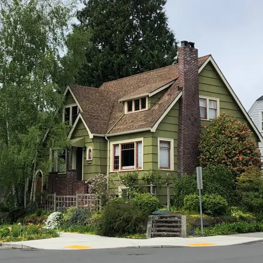 A country house design with olive green siding and a prominent brick chimney, surrounded by mature trees.