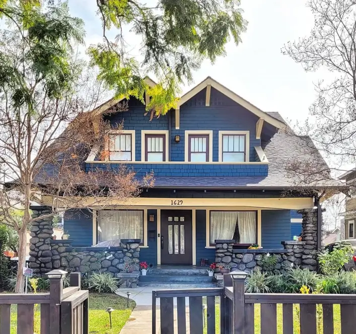 A deep blue craftsman house exterior with a wide porch featuring river rock columns and a prominent dormer window.