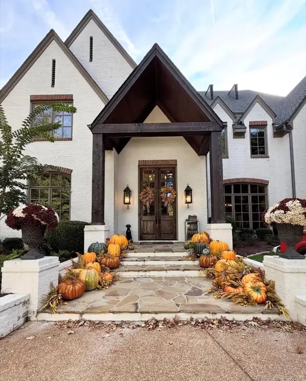An off-white European-style farmhouse with a dark wood entrance and stone steps decorated with pumpkins.
