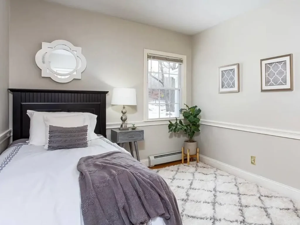 A small, cozy bedroom with a dark wood bed, neutral gray walls with chair rail molding, and a plush diamond-patterned rug.