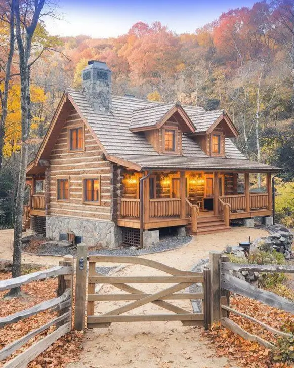 Small Log Cabins: 36 Ideas for Cozy Living & Enduring Charm 27 A two-story small log cabin house with a stone foundation and glowing porch lights in an autumn forest.