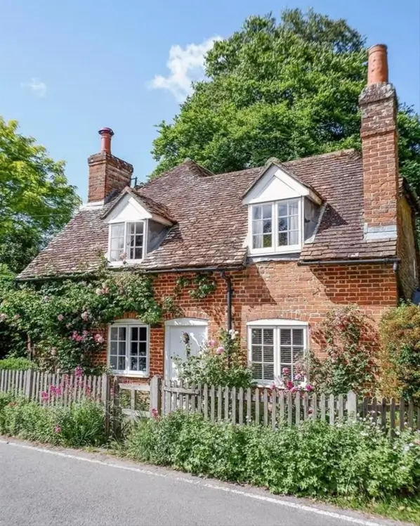 A classic British country house made of red brick with climbing pink roses and a wooden picket fence.