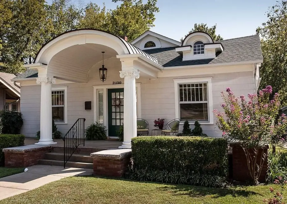 An off-white craftsman bungalow with a unique arched portico, classical white columns, and a dark green front door.