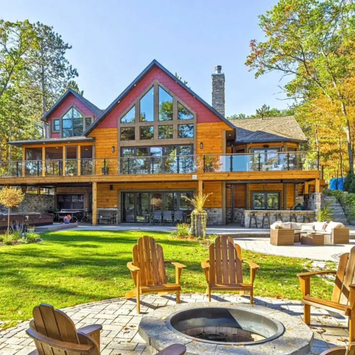 Embracing Grandeur: 25 Inspiring Ideas for Large Log Cabin Homes 28 A modern log cabin with glass deck railings, a lower patio with an outdoor kitchen, and a fire pit in the foreground.