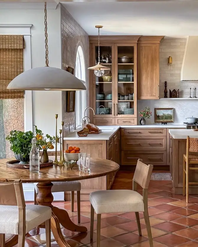 A rustic kitchen design idea with warm terracotta floor tiles, natural wood cabinets, and a round wooden dining table.