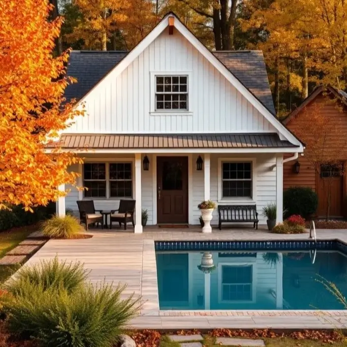 A white farmhouse-style cottage with a poolside deck, surrounded by lush greenery and vibrant orange trees in the fall.
