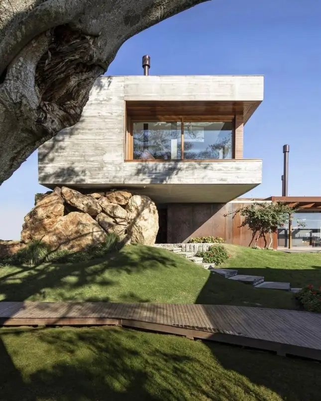 A cantilevered modern concrete home balanced on a large natural boulder, with a wood-framed window.