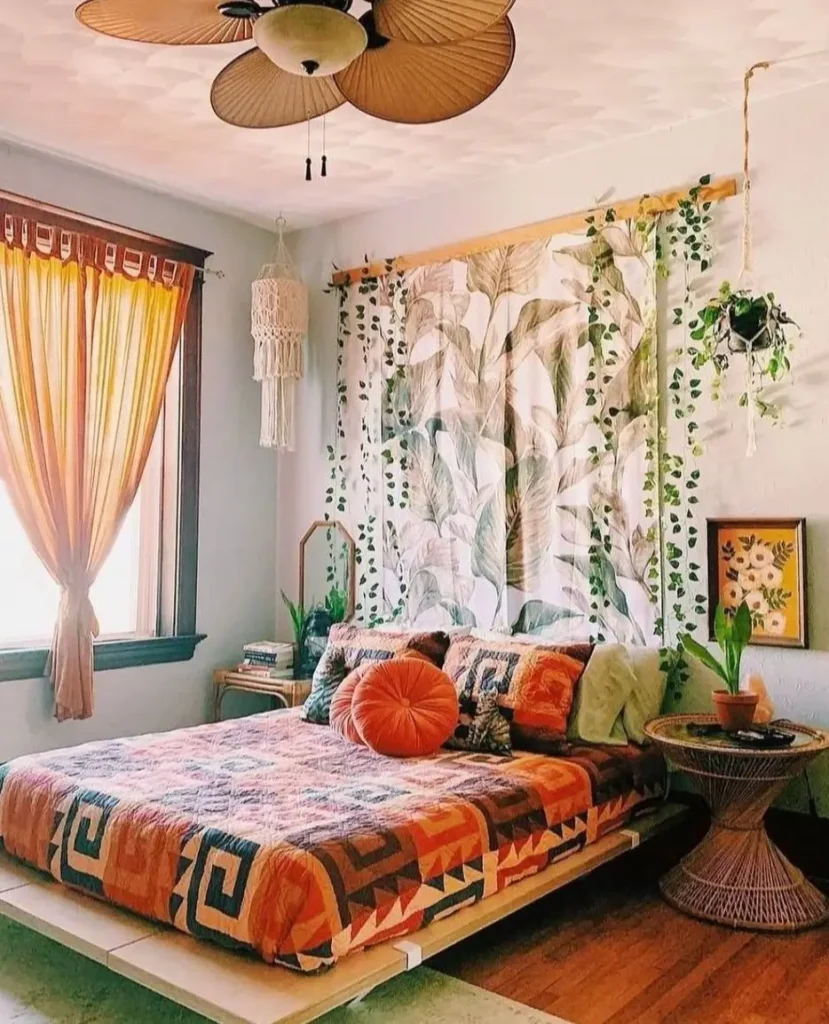 A boho bedroom featuring a large botanical leaf-print tapestry hanging on the wall behind a low platform bed, with ivy and a macrame hanger nearby.
