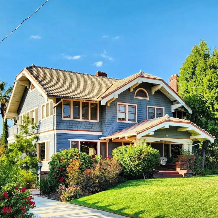 A large craftsman house exterior with dusty blue siding and a warm terracotta tile roof, surrounded by lush landscaping.