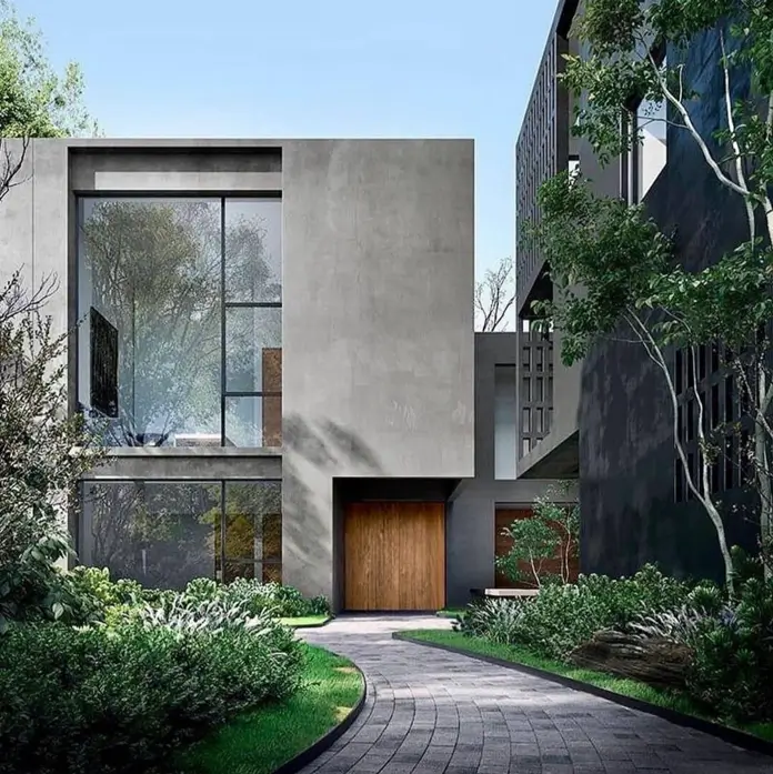 A modern concrete home with a large window and wooden door, surrounded by lush gardens and a stone path.