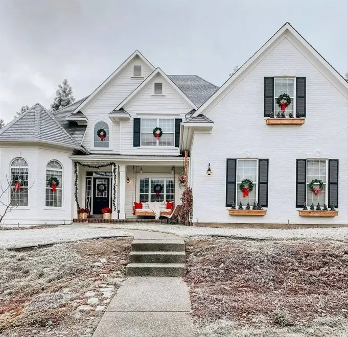 A beautiful off-white brick farmhouse with black shutters and window boxes, decorated for winter with a light frost on the ground.