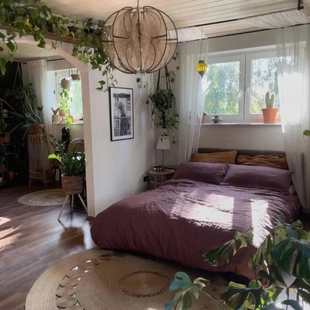 A sun-drenched bedroom with a bed dressed in deep plum-colored linens, surrounded by plants and natural light.
