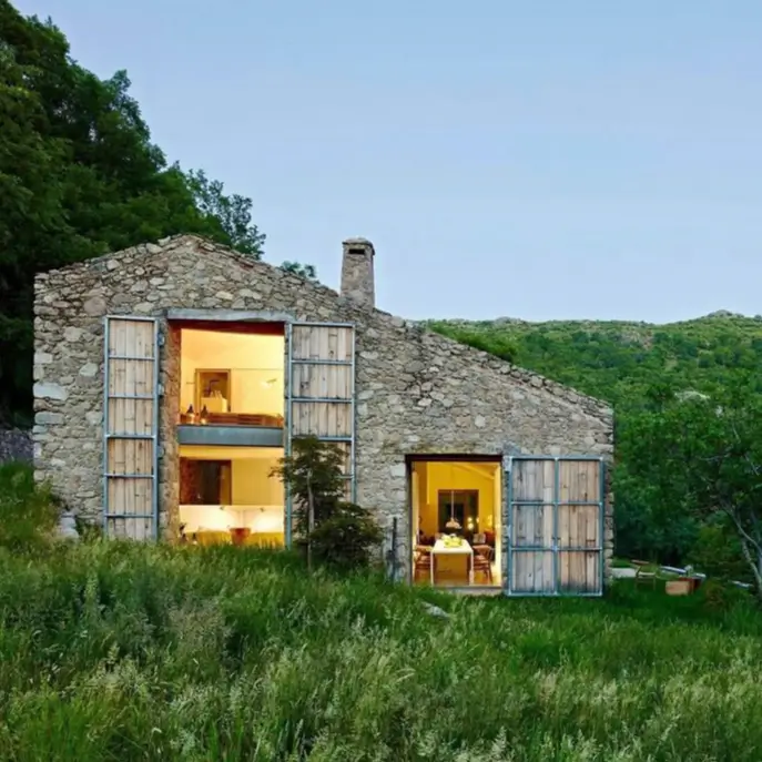 A modern country house design within a rustic stone barn, featuring large openings and wooden shutters at dusk.