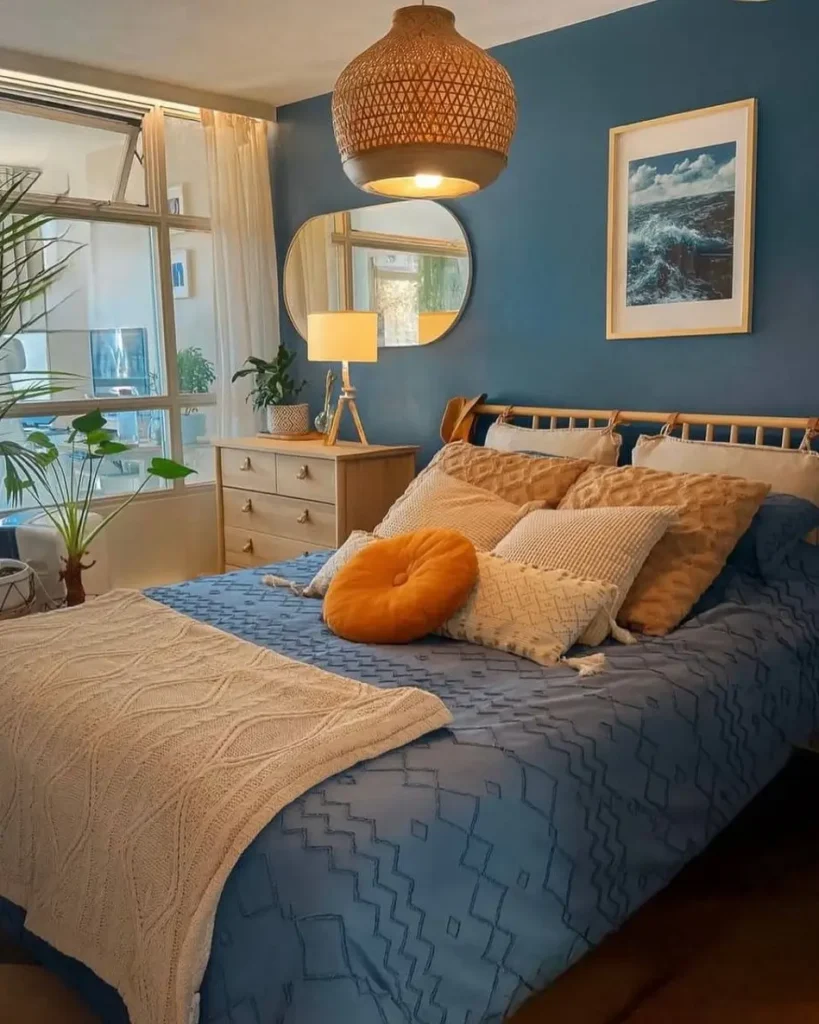 A cozy bedroom with deep blue walls, a bed with blue bedding, and a variety of warm, textured pillows.