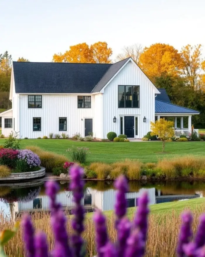 A modern white farmhouse with a pond, viewed through out-of-focus purple flowers, with golden autumn trees in the background.