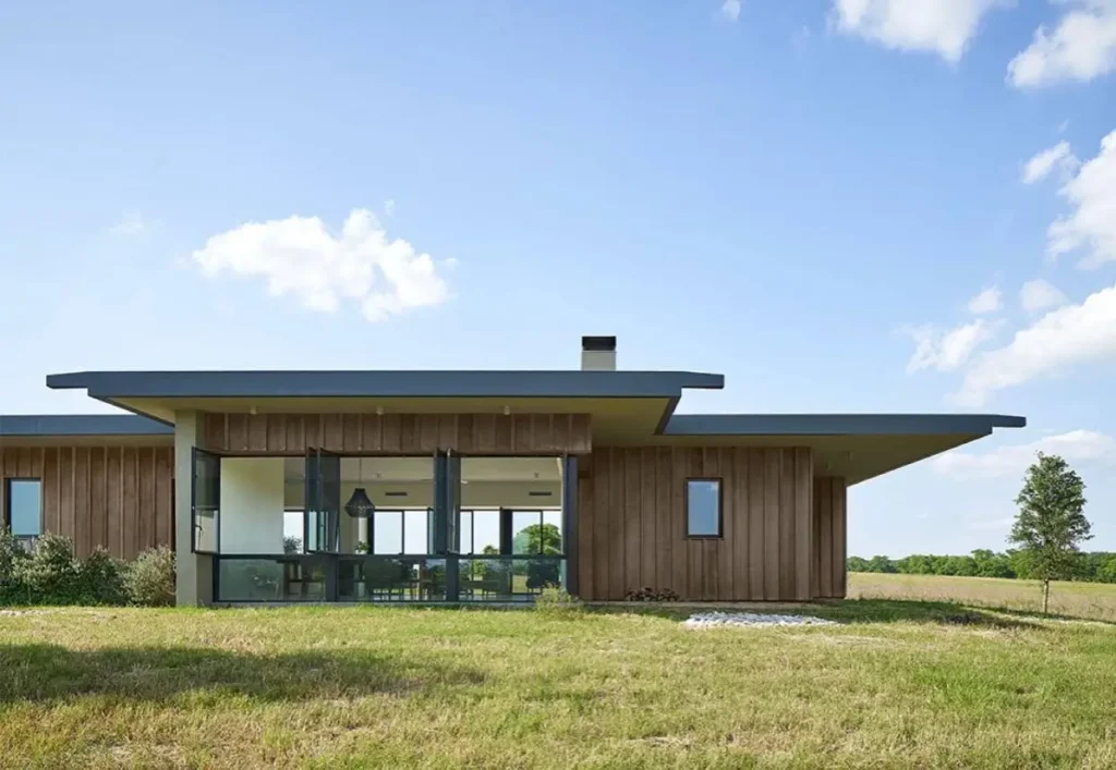 An architectural ranch house with a prominent extended roofline and vertical wood siding in a field.