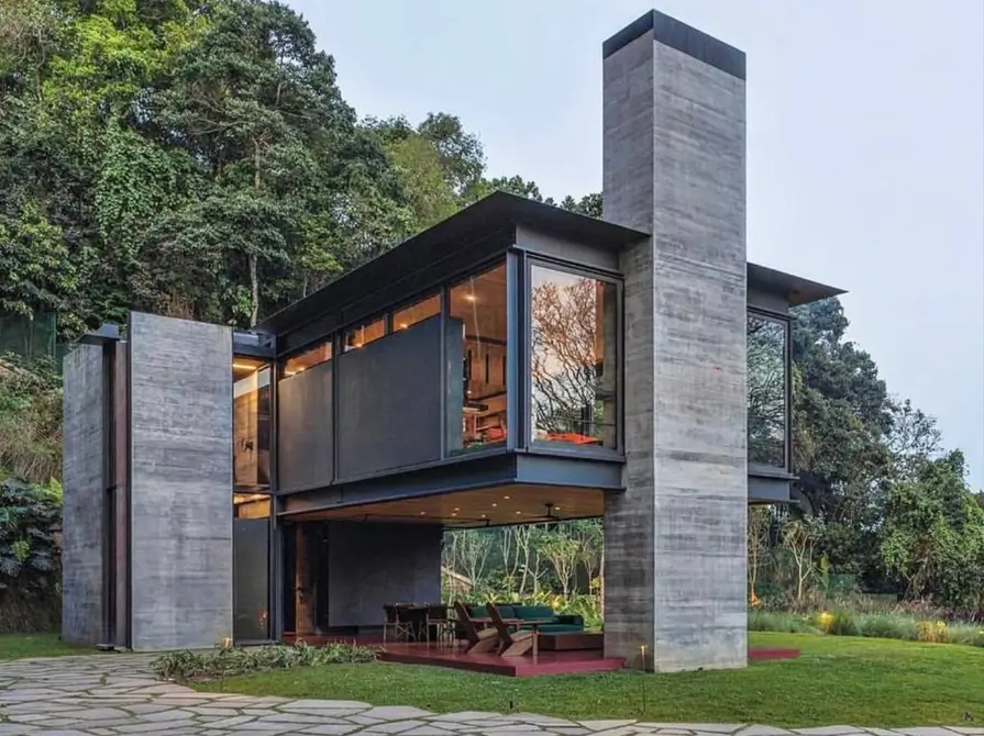 A modern concrete home nestled in a forest, featuring large glass walls and a cantilevered second floor.
