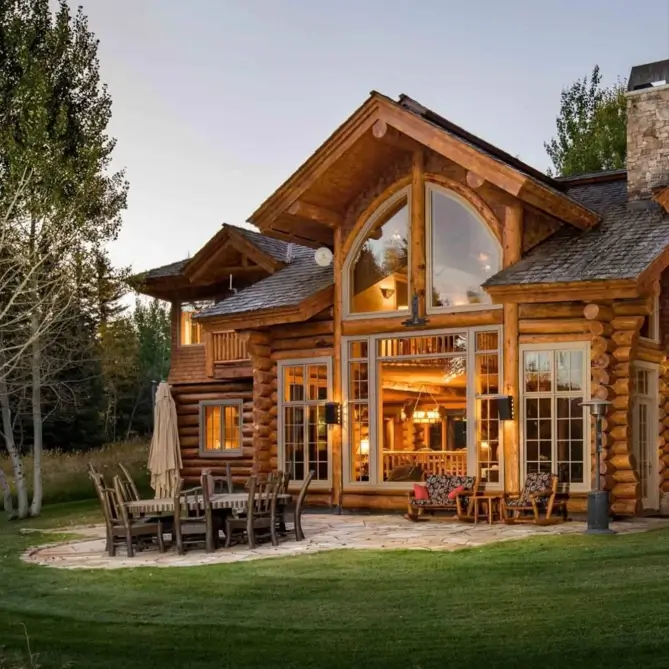 Embracing Grandeur: 25 Inspiring Ideas for Large Log Cabin Homes 6 Large log cabin home at dusk with a warmly lit interior visible through an arched window and a furnished stone patio.