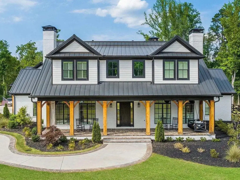 A two-story modern white farmhouse exterior with a striking dark metal roof, black-trimmed windows, and a deep front porch with light wood support beams.