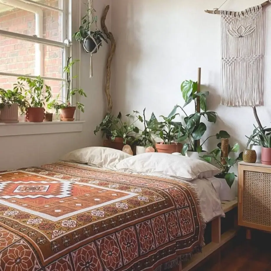 A sunlit bedroom with a low wooden bed covered by a terracotta patterned bohemian throw, surrounded by houseplants.