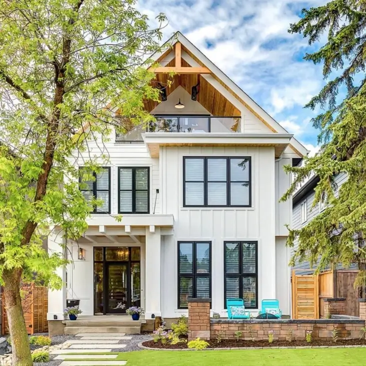 A modern off-white farmhouse featuring an exposed wood truss in the highest gable and large black windows.