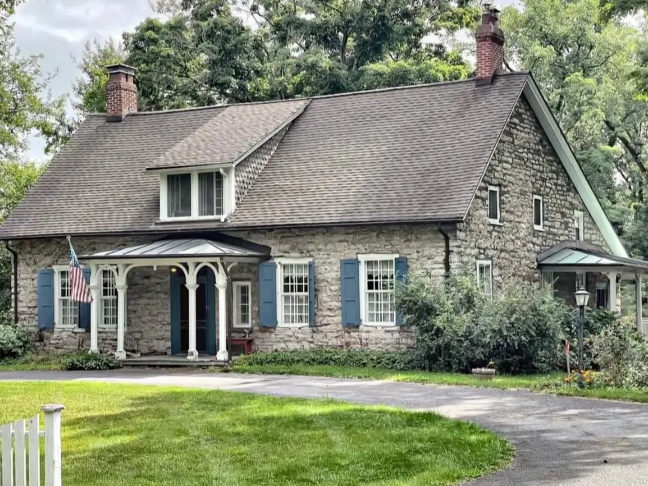 A large stone cottage home with blue shutters, a white front porch, and a driveway.