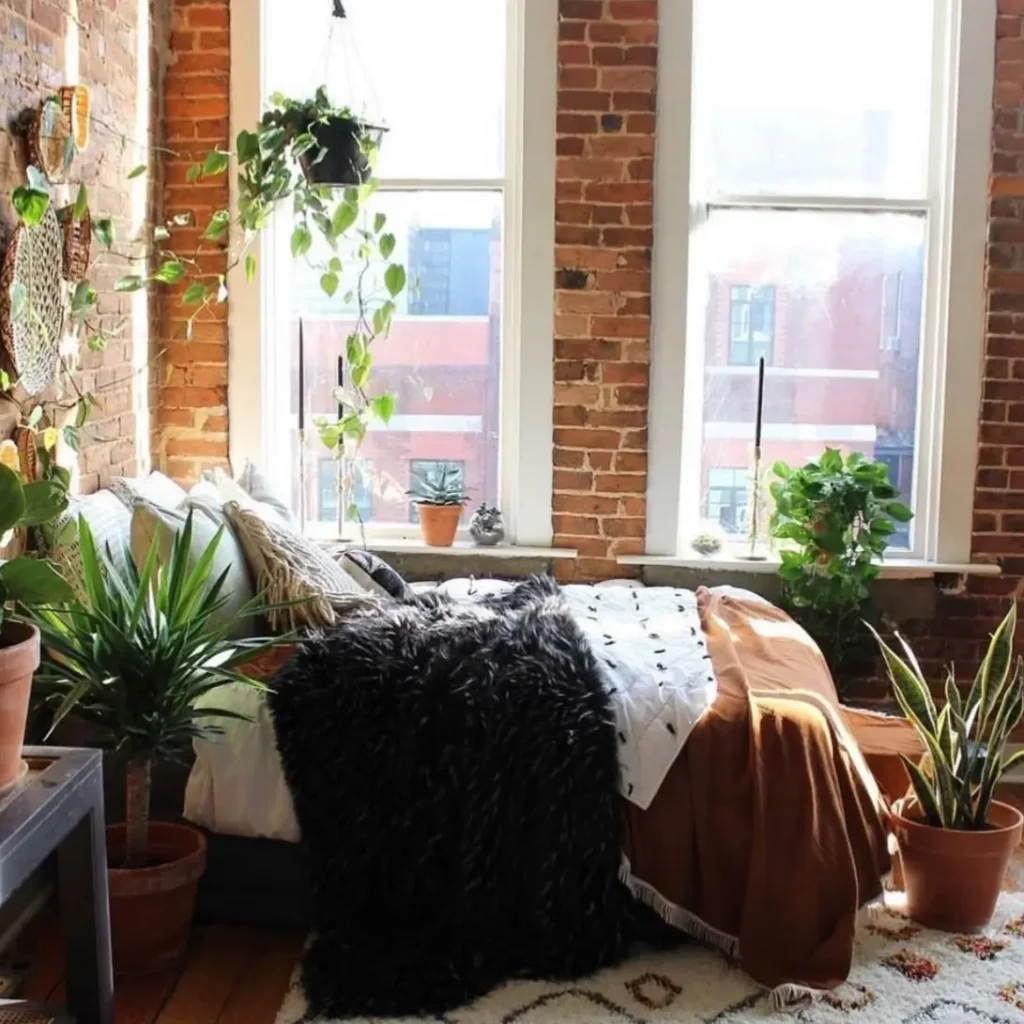 A sunlit bedroom with exposed brick walls, filled with numerous houseplants and a cozy bed layered with textured blankets.