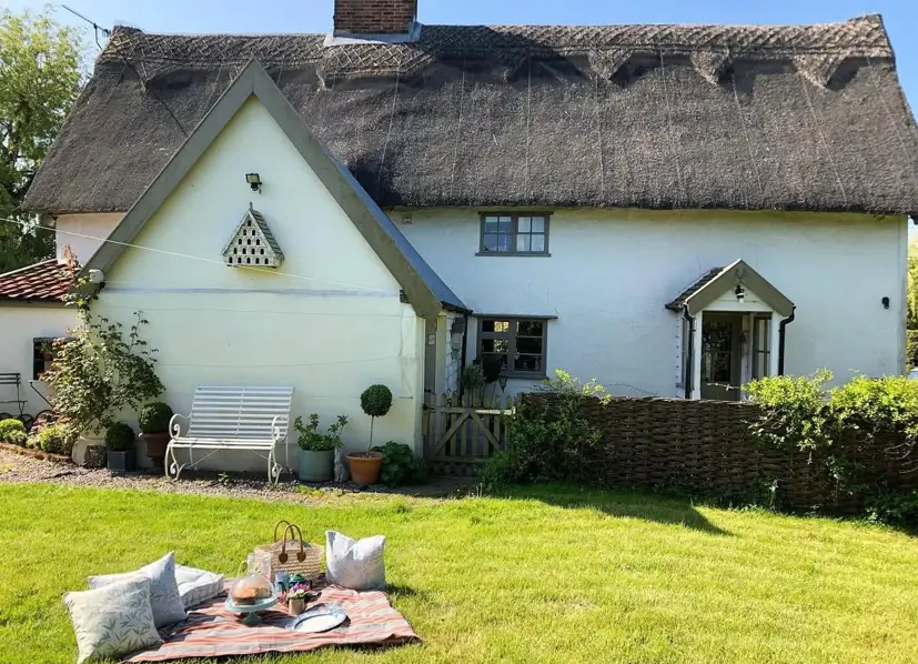 A white thatched British country house with a woven wattle fence and a picnic set up on the lawn.