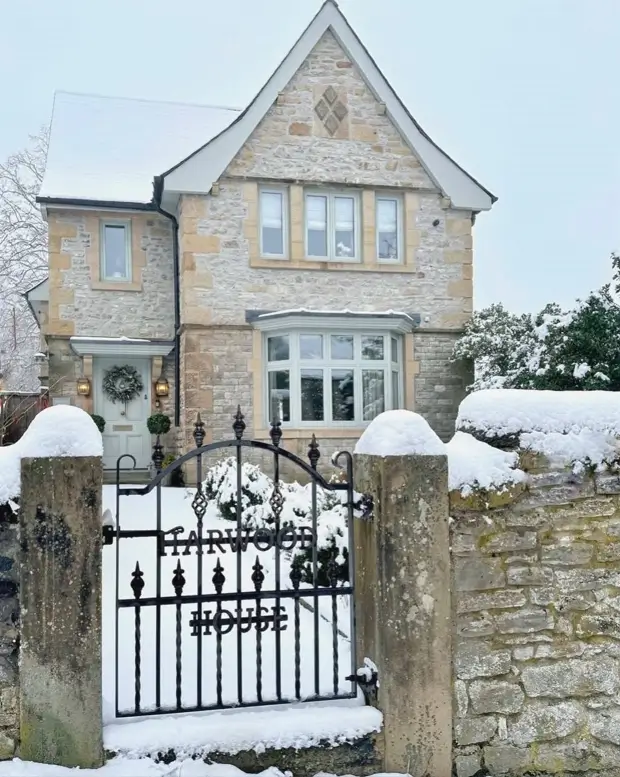 A stone English country house in the snow behind a black wrought iron gate and a low stone wall.