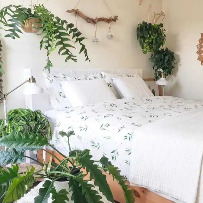 A bright bedroom with white bedding featuring a green leaf pattern, surrounded by lush hanging and potted plants.