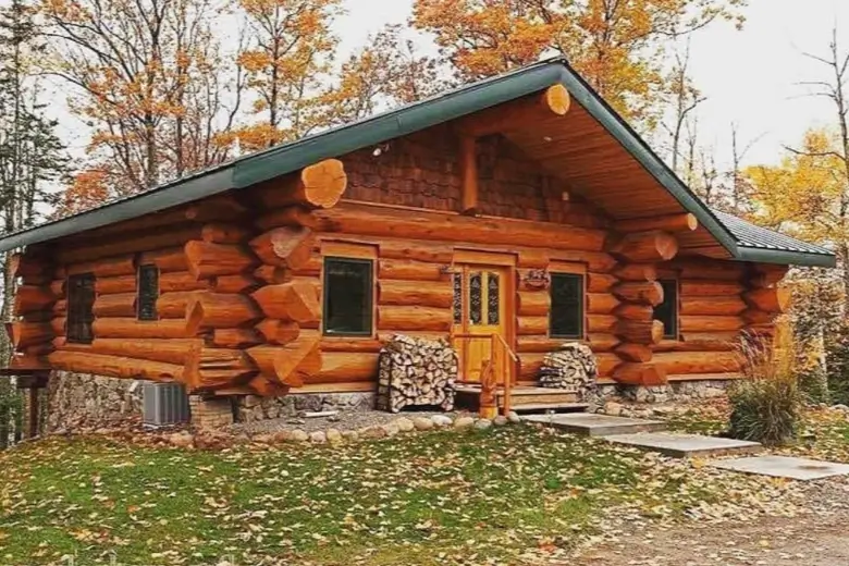 60 Charming Small Log Cabin Ideas to Inspire Your Rustic Retreat 33 A classic small log cabin home with a stone foundation and green roof, set against a backdrop of autumn trees.