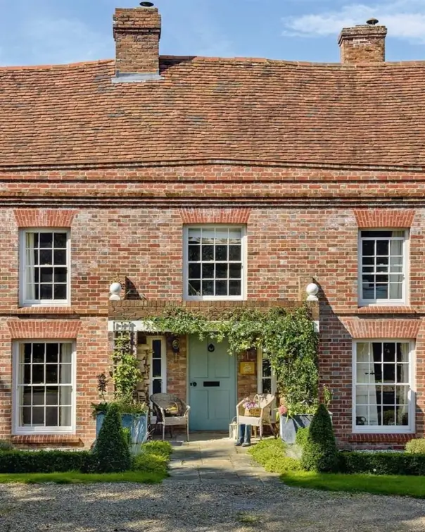 A red brick British country house with a blue door and a sheltered entryway framed by chairs and climbing plants.
