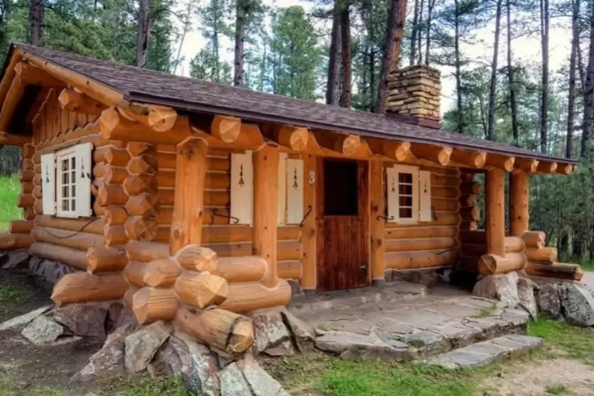 60 Charming Small Log Cabin Ideas to Inspire Your Rustic Retreat 34 A rustic small log cabin home with a stone foundation, white shutters, and a stone chimney in a pine forest.