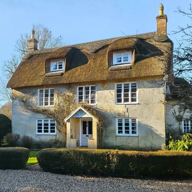 A classic country house design featuring a traditional thatched roof and weathered stucco walls in the bright sun.